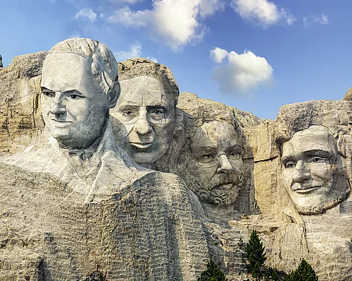 Vier Männergesichter als Felsrelief im Stil von Mount Rushmore bei blauem Himmel